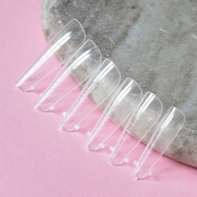 Seis unhas postiças transparentes em superfície cinzenta sobre fundo rosa