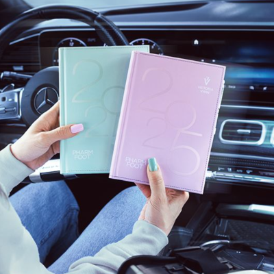 Dois cadernos pastel azul e rosa com relevo e texto, segurados dentro de um carro moderno.