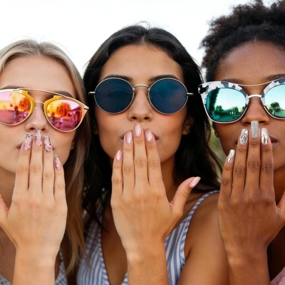Três mulheres com óculos de sol coloridos e unhas decoradas, mostrando as mãos perto da face.