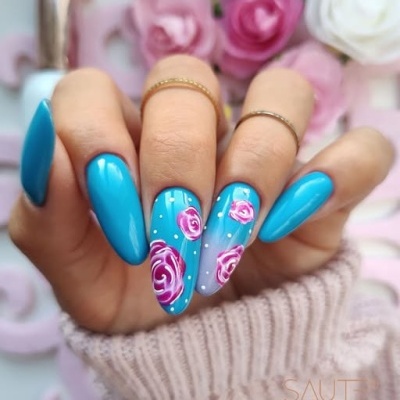 Mão com unhas azuis decoradas com flores rosas e bolinhas brancas, anéis dourados finos e flores ao fundo