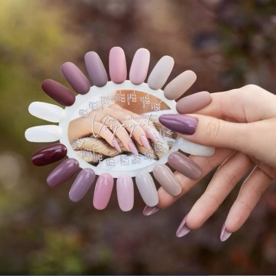 Mostruário circular de unhas postiças em tons pastel, nude e roxos, com mão segurando.