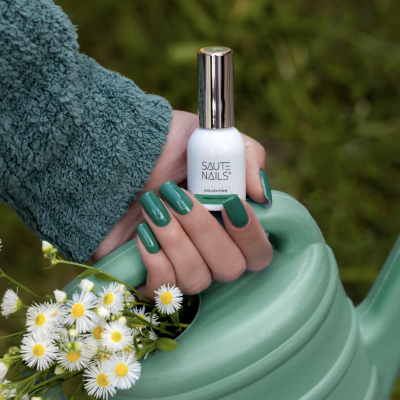 Frasco de verniz branco com tampa dourada numa mão com unhas verdes junto a regador e flores