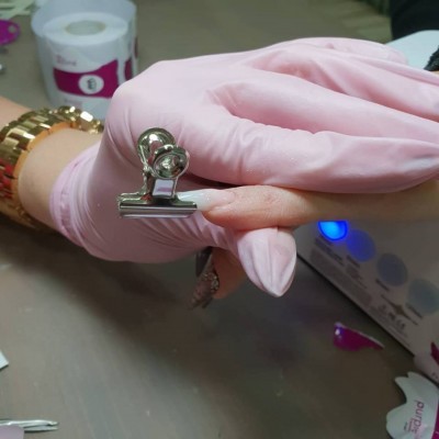 Mãos com luvas cor-de-rosa segurando clips metálico em unha com gel transparente e luz azul de aparelho para unhas