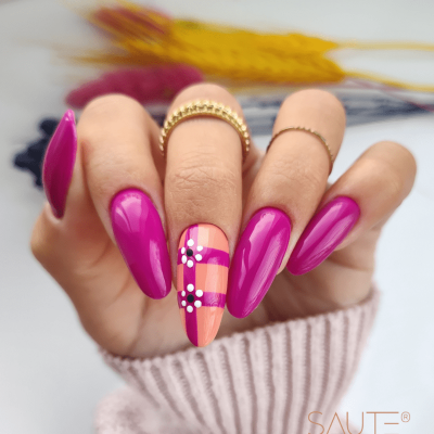 Unhas cor rosa brilhante com decoração xadrez e flores, anéis dourados e fundo com plantas secas desfocadas