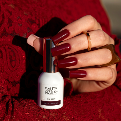 Frasco de verniz para unhas SAUTE NAILS DEEP CHERRY com mão segurando e fundo vermelho de renda.