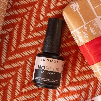 Frasco de verniz para unhas INOCOS NO BLUE TOP COAT sobre tecido laranja padrão geométrico ao lado de embrulho com fita vermelha