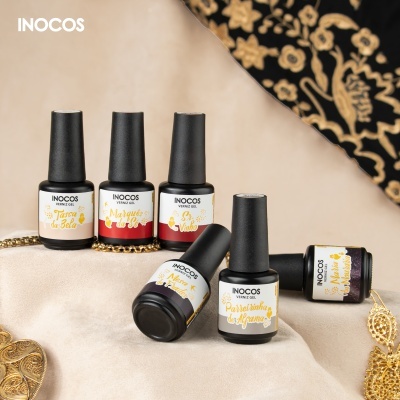 Frascos de verniz gel INOCOS sobre tecido bege claro com objetos dourados e tecido preto com padrão dourado.