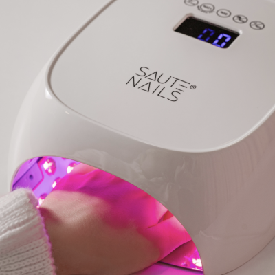 Dispositivo branco para unhas com visor digital azul e luzes LED rosas a iluminar a mão de uma pessoa.