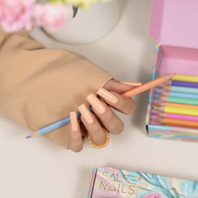 Mão com unhas nude segurando lápis de cor azul claro ao lado de caixa de lápis coloridos e caixa de verniz para unhas em fundo branco