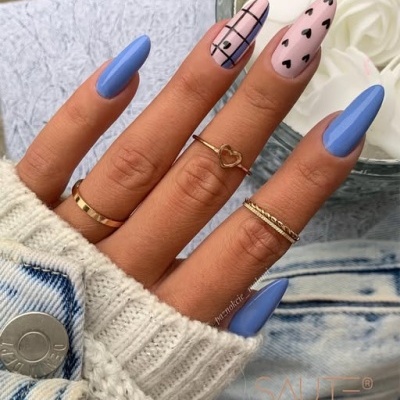 Mão com unhas pintadas de azul e rosa com padrões, anéis dourados e flores brancas ao fundo.