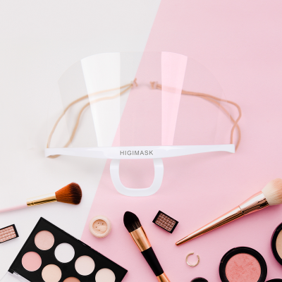 Máscara facial transparente HIGIMASK com suporte branco e cordão elástico bege entre produtos de maquilhagem sobre fundo branco e rosa