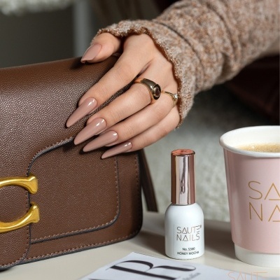 Mão com unhas nude segurando mala castanha ao lado de verniz branco e copo rosa SAUTE NAILS