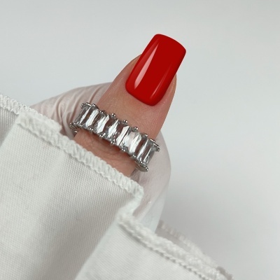 Anel prateado com pedras retangulares no dedo polegar de mão com unha vermelha, fundo branco.