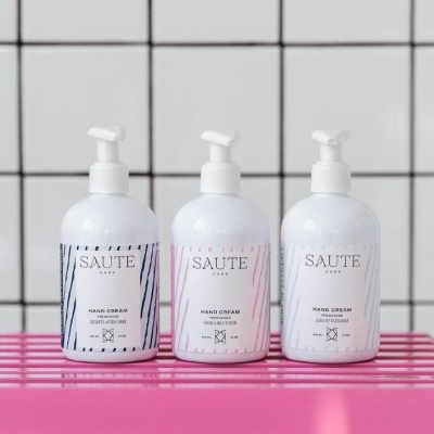 Três frascos de creme para as mãos SAUTE CARE com dispensador em superfície rosa diante de parede de azulejos brancos.