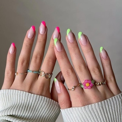 Mãos femininas com unhas decoradas em rosa neon e verde neon com base nude, usando anéis delicados, fundo cinzento.