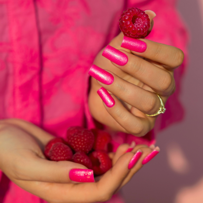 Mãos femininas com unhas cor de rosa brilhante segurando framboesas, com roupa rosa ao fundo