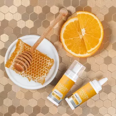 Produtos em frascos amarelos e brancos, favo de mel num prato branco e fatia de laranja sobre padrão hexagonal bege.