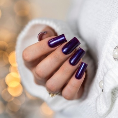 Unhas artificiais roxas brilhantes em mão feminina com fundo desfocado e roupa branca.