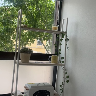 Estante branca de metal com prateleiras contendo planta, caneca, velas, toalhas e aparelho, próxima a janela e parede branca