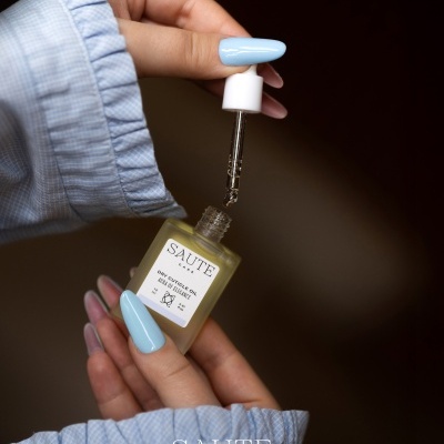 Frasco de óleo para cutículas seco SAUTE CARE com conta-gotas a ser segurado por mãos femininas com unhas azuis claras.