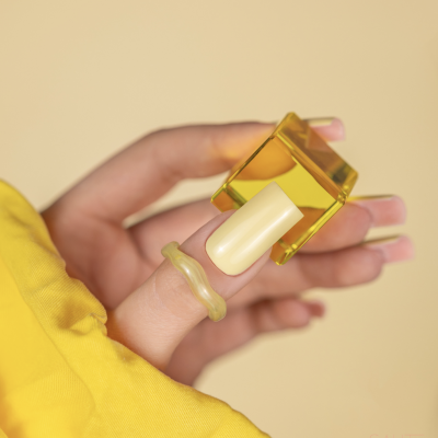 Mão com unhas manicadas segurando dois anéis amarelos translúcidos, fundo e roupa amarelo claro.