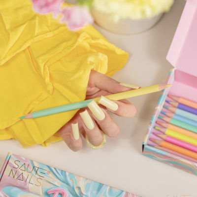 Mão com unhas amarelas segurando lápis de cor pastel sobre caixa aberta de lápis coloridos pastel da marca SAUTE NAILS Pastel.