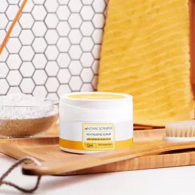 Frasco branco de esfoliante revitalizante com toalha amarela e escova sobre tabuleiro de madeira