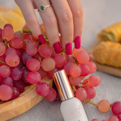 Frasco de verniz SAUTE NAILS e mão com unhas vermelhas junto a uvas rosadas