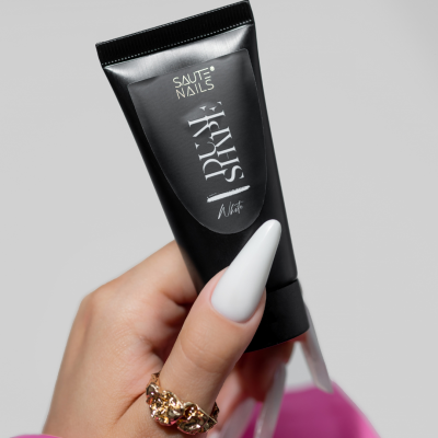 Mão com unhas brancas segurando tubo preto de cosmético SAUTE NAILS, fundo cinza