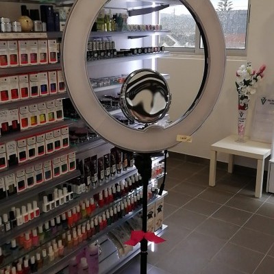 Anel de luz branco com espelho no centro em tripé preto junto a estantes com vernizes e cosméticos em sala com janela e chão cinza