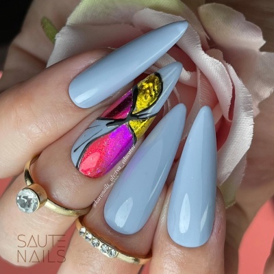 Unhas azul claro e decoradas com padrão fractal colorido, com anéis dourados e flor cor de rosa