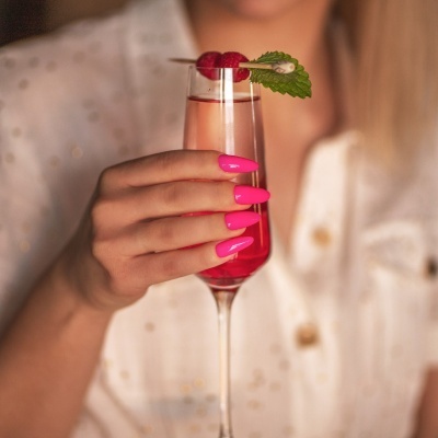 Taça de vidro com bebida vermelha decorada com framboesas e uma folha verde, mão com unhas cor de rosa e camisa branca