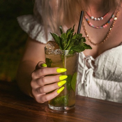 Copo com bebida e hortelã na mão de pessoa com unhas amarelas florescentes
