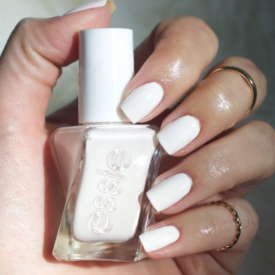 Frasco de verniz Essie branco opaco numa mão com unhas brancas e anéis dourados