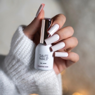 Frasco de verniz branco Saute Nails com unhas brancas numa mão e fundo desfocado