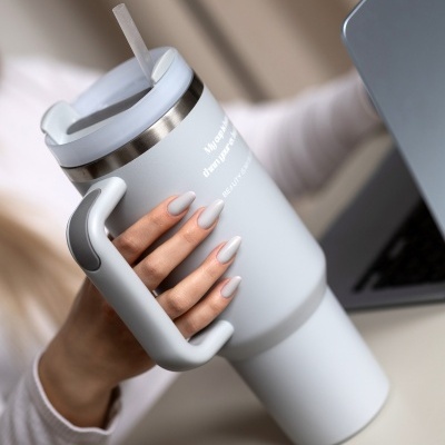 Caneca térmica cinzenta clara com pega e texto branco, mão com unhas bege mate segurando-a junto a portátil.