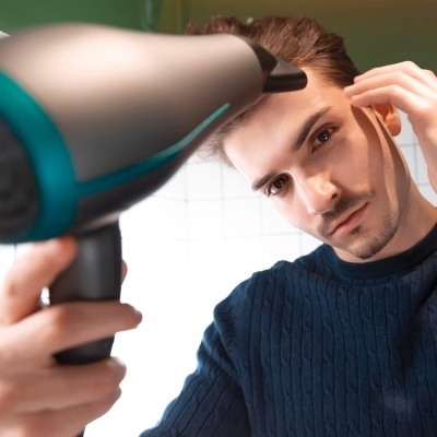 Homem a usar secador de cabelo preto e azul, vestindo suéter azul escuro em frente a azulejos brancos