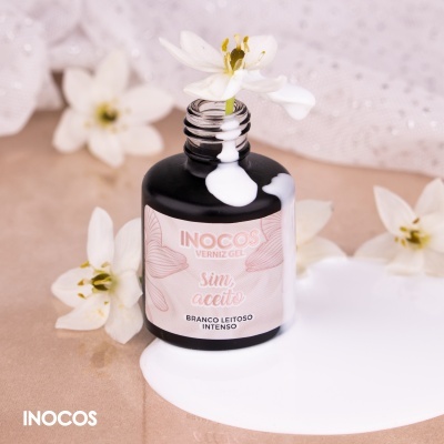Frasco preto de verniz gel INOCOS branco leitoso com verniz derramado e flores brancas ao redor