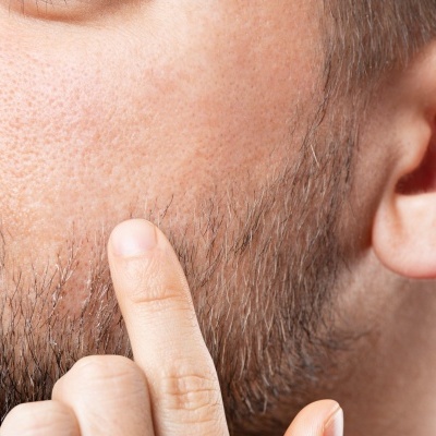 Close-up do lado esquerdo do rosto de um homem com barba castanha e dedo tocando a barba