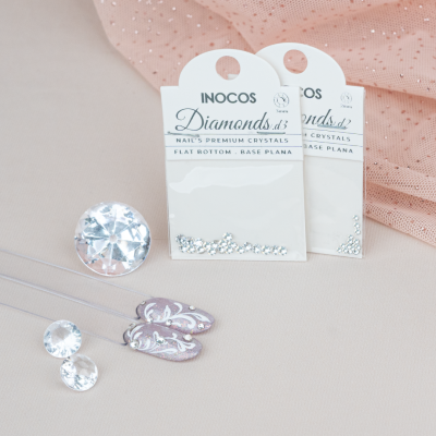 Cristais para unhas INOCOS Diamonds em embalagem branca sobre tecido rosado brilhante com unhas decoradas lilás.