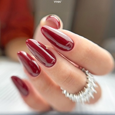 Unhas vermelhas brilhantes com anel prateado e logótipo VICTORIA VYNN ao fundo