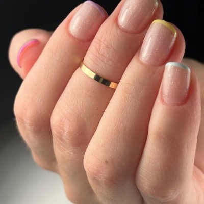 Mão com unhas pintadas de esmalte transparente e bordas coloridas, e anel dourado simples