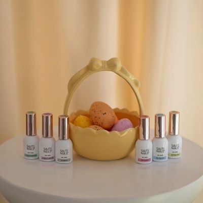 Vernizes SAUTE NAILS brancos com tampas douradas ao redor de um cesto amarelo com ovos coloridos com manchas pretas.