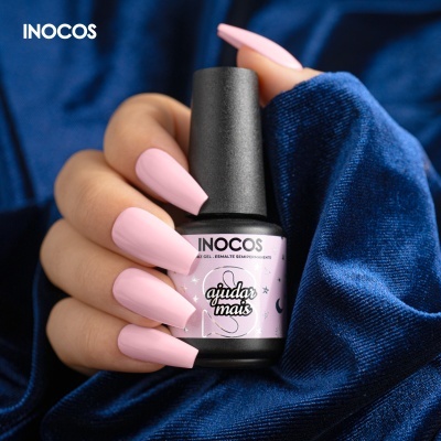 Mão com unhas rosa claro segurando verniz gel INOCOS com fundo azul escuro aveludado
