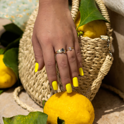 Mão feminina com unhas amarelas tocando limão junto a bolsa de palha e mais limões
