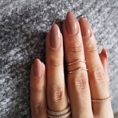 Mão feminina com unhas nude e anéis dourados e prateados sob fundo cinzento