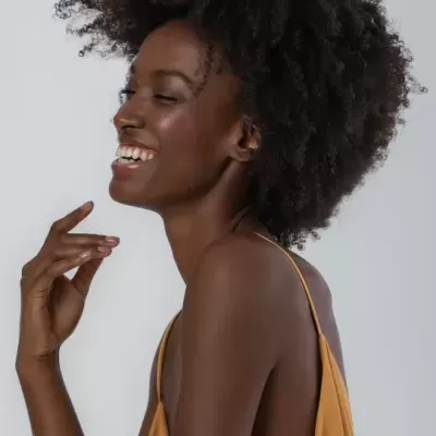Mulher negra com vestido castanho claro de alças finas a sorrir e de perfil