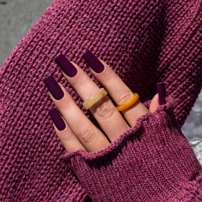 Mão com unhas roxas e anéis em resina sobre casaco de malha roxa