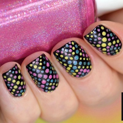 Unhas pintadas de preto com bolinhas coloridas, segurando frasco de verniz rosa holográfico