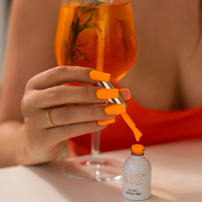 Mão com unhas laranja a pintar com verniz SAUTE NAILS junto a cocktail laranja